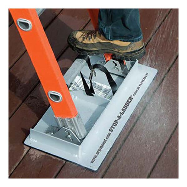 A person in work boots is standing on an orange ladder, which is securely held in place on a wooden deck by the Working Concepts Stop-A-Ladder 7002. This device, designed by Working Concepts, enhances extension ladder safety and prevents slipping.